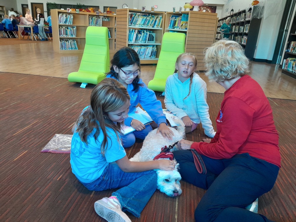 three fourth grade girls pet a small white dog wearing a red collar, as a woman wearing blue pants and a red sweater speaks to one of them