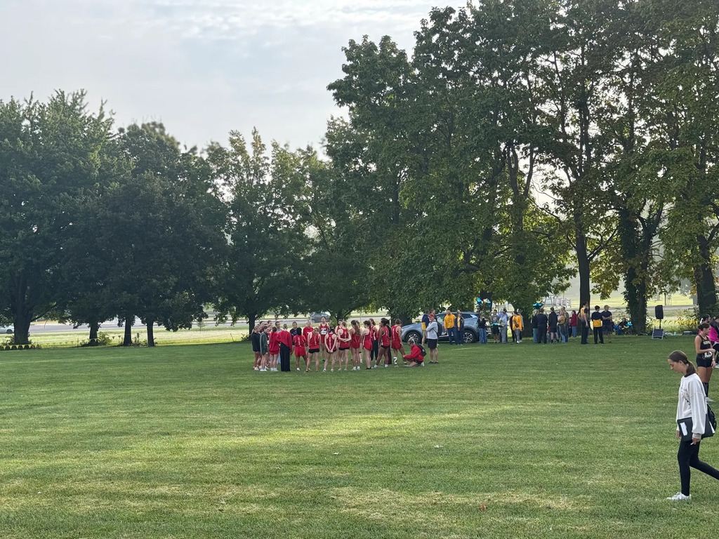 runners wearing red uniforms stand in a circle in a grassy field with trees in the background