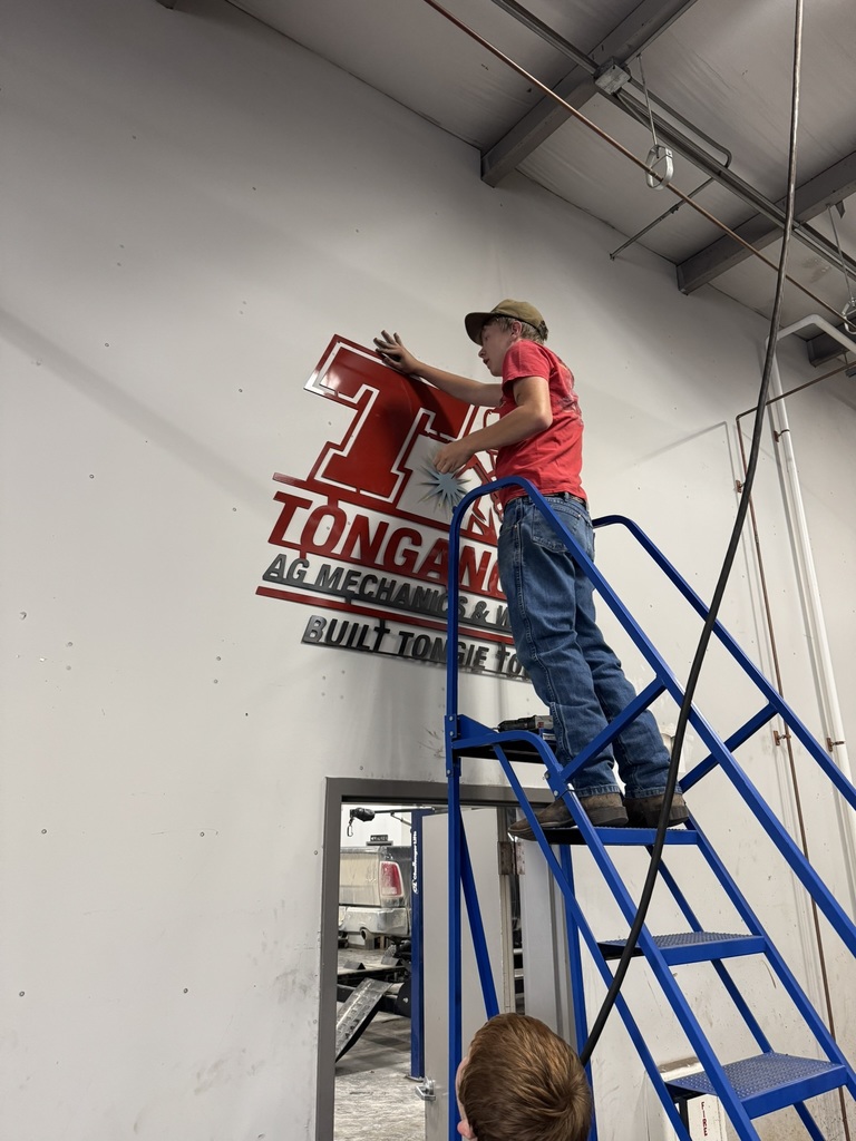 a student wearing jeans and a red shirt, secures a welded sign to the wall above the shop entrance