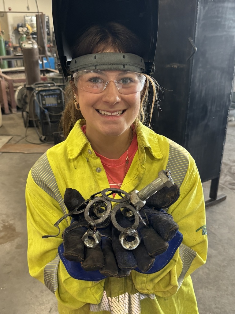 Lilly Bedtke wears a yellow welder's jacket, gloves, glasses, and hood pulled back to reveal her face; she holds a welded creation made out of bolts and screws