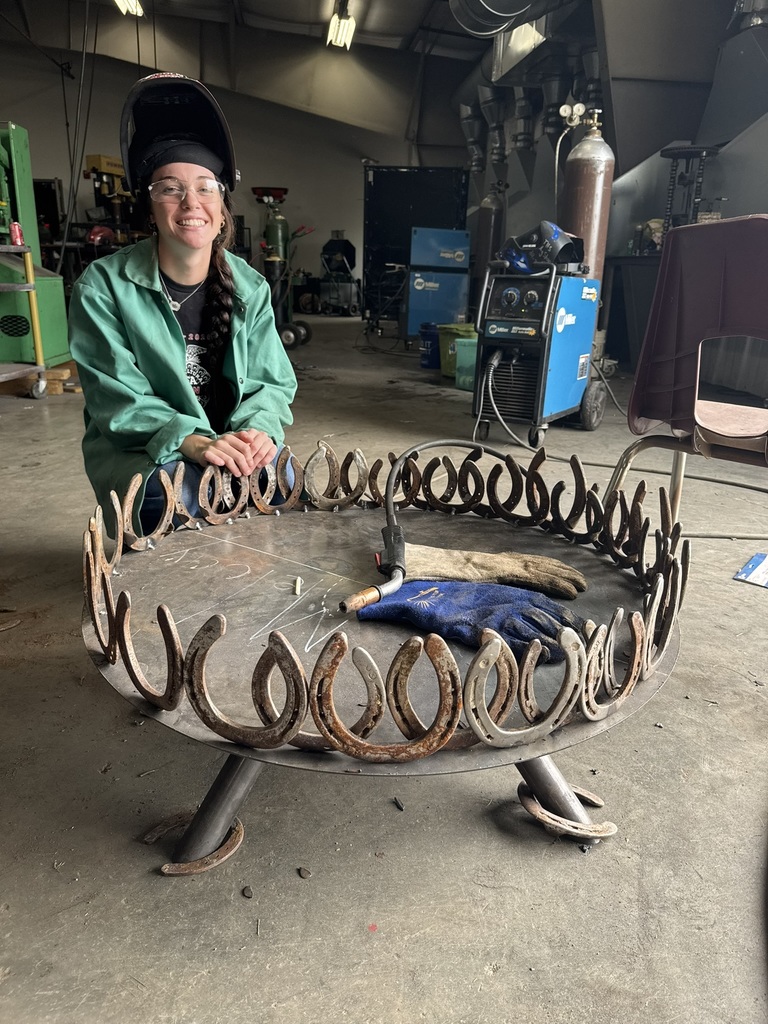 a welding student kneels down to pose by her horseshoe fire pit