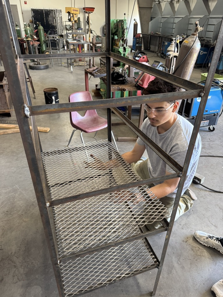 a studetn wearing khakis, a gray t shirt, and safety glasses puts together a metal shelving unit he built