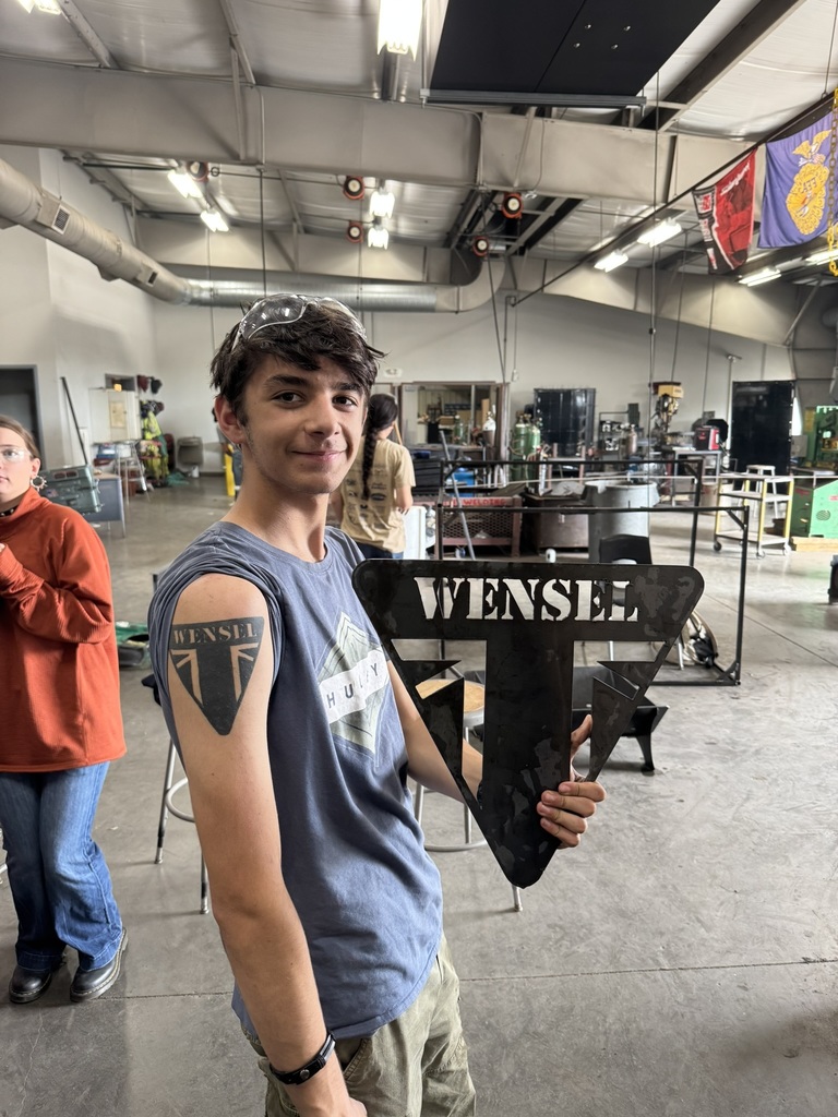 student wearing a blue shirt, holding an upside down triangle shaped metal shield with the word WENSEL on it; a matching tattoo can be seen on his bicep
