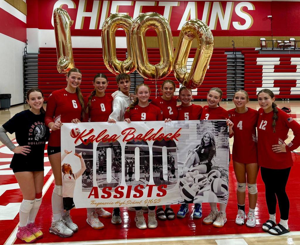 kalea baldock wearing a red volleyball uniform, flanked by team members on a volleyball court. they hold up a banner that says kalea baldock 1000 assists tonganoxie high school 9.16.25