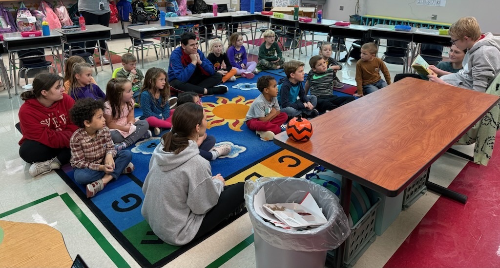 Kindergarten students sitting on the carpet listening to a high school student read to them. 