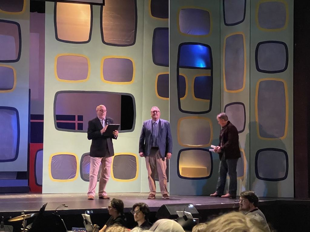 Mr Harrell stands on a stage, flanked by a man holding a microphone and a man holding an award