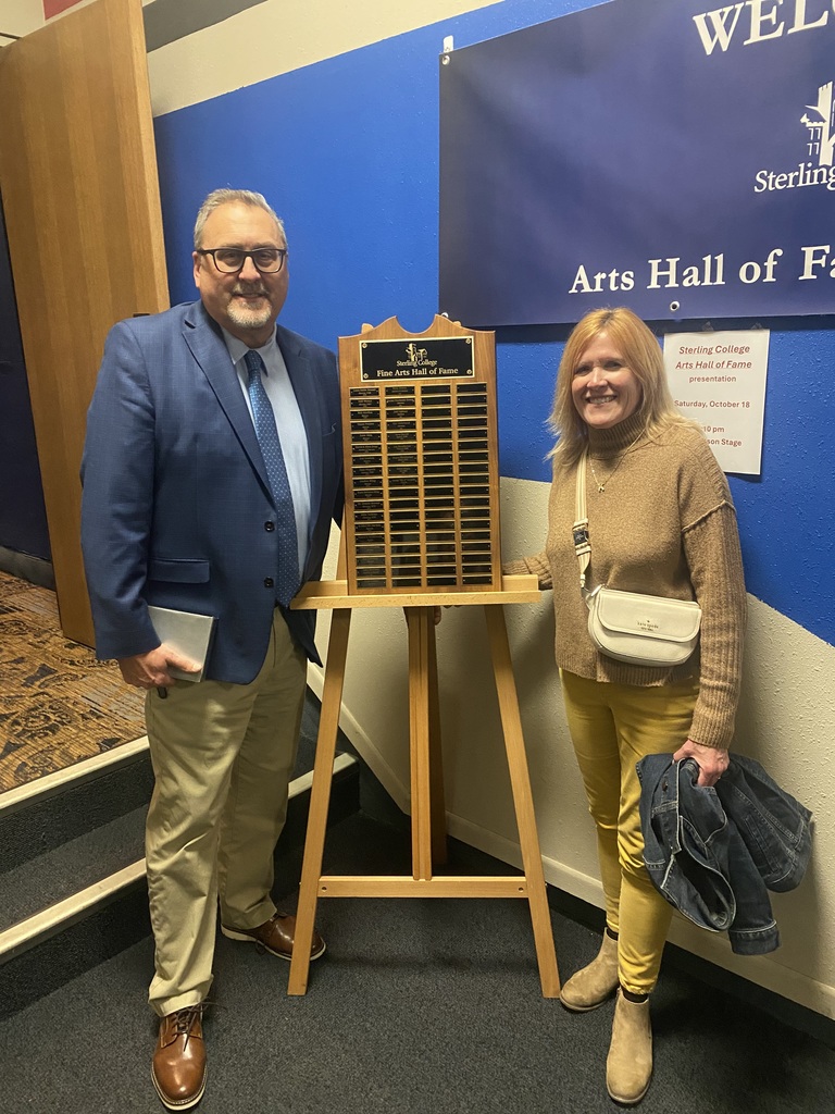 Mr Harrell wearing glasses, a light blue shirt, blue tie and sport coat, and khakis; Mrs. Harrell wearing a brown sweater and yellow pants; a wooden easel with metal name plaques stands between them