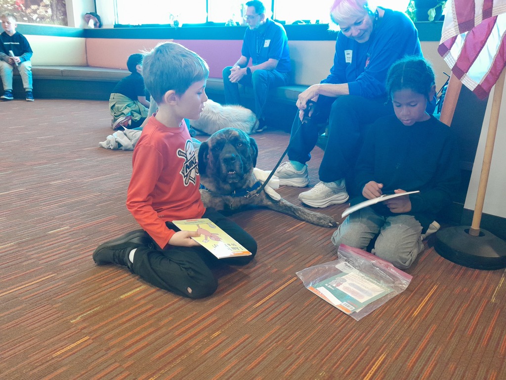 third graders sitting on a carpeted floor with a brown dog wearing a blue leash
