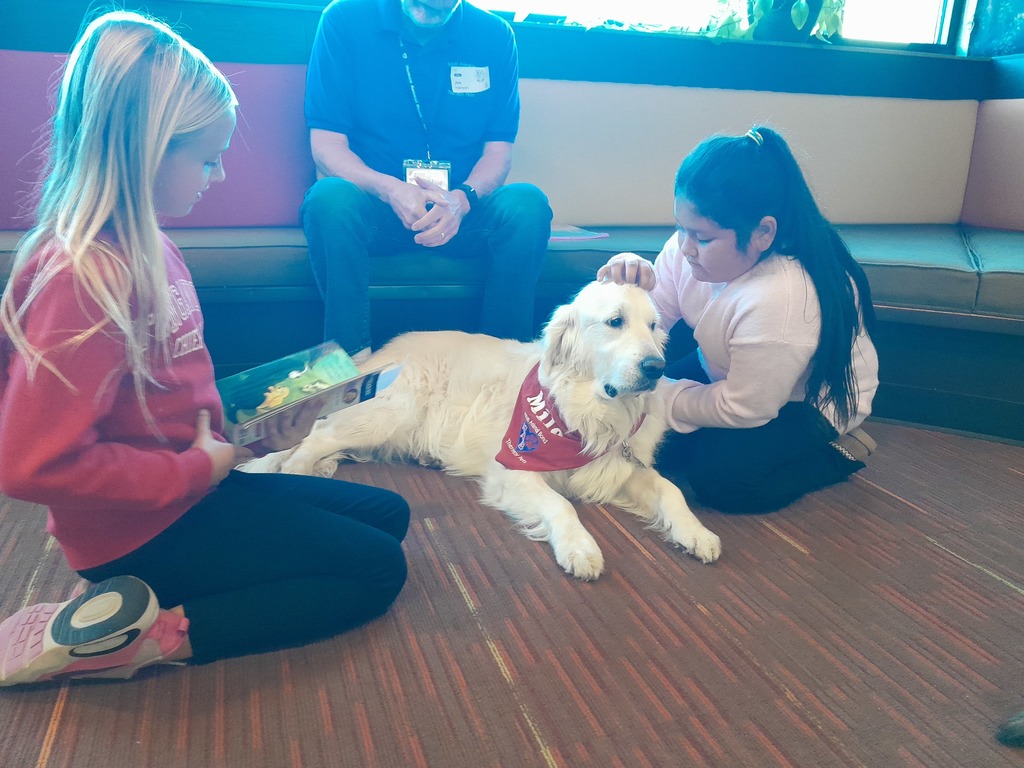 third graders sitting on a carpeted floor with a white dog wearing a red bandanna