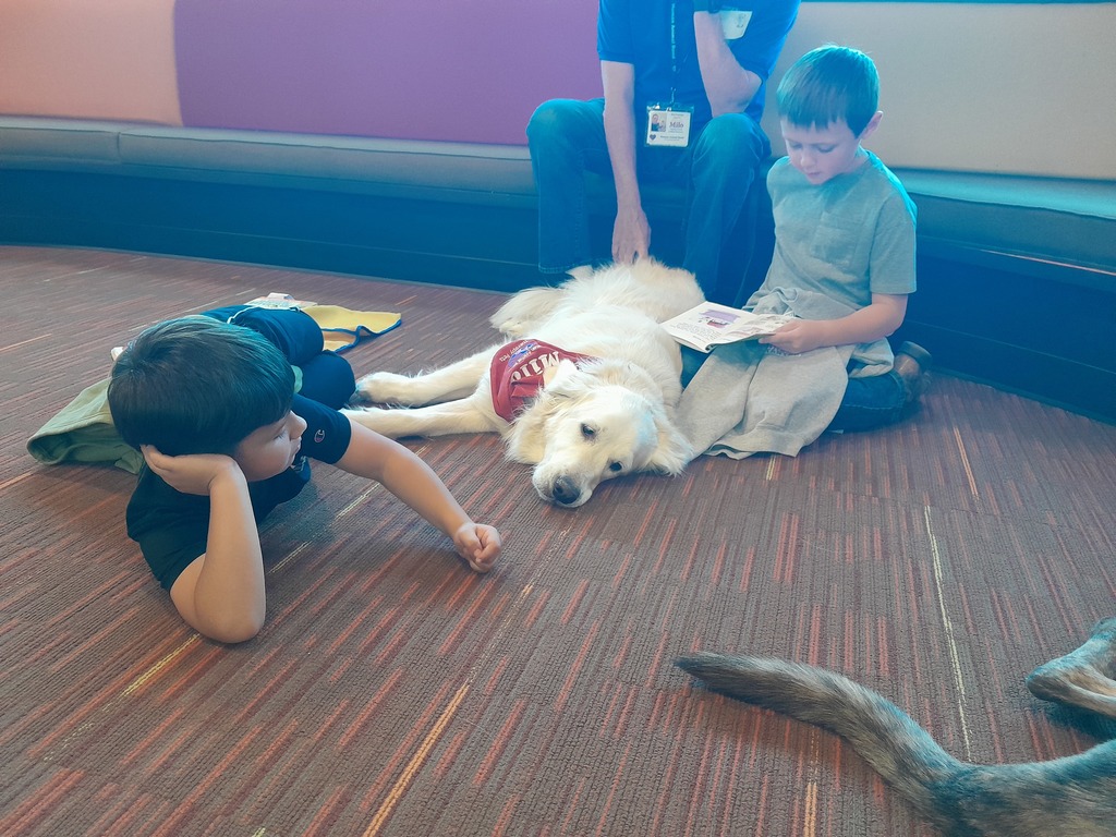 third graders sitting on a carpeted floor with a white dog wearing a red bandanna