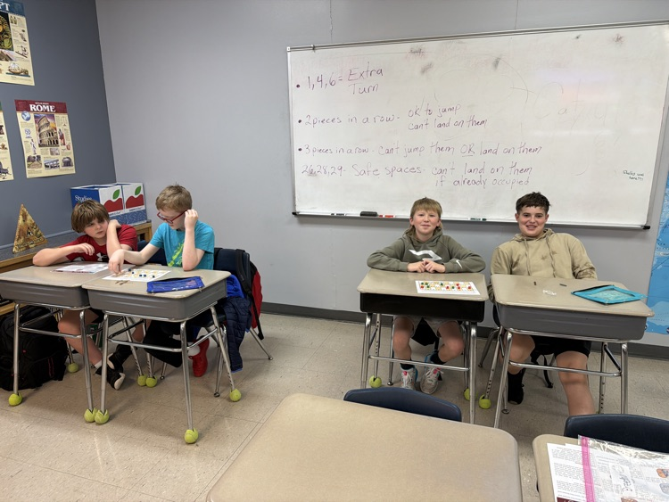 four sixth grade students sit at gray school desks; a whiteboard hangs on a light blue wall behind them