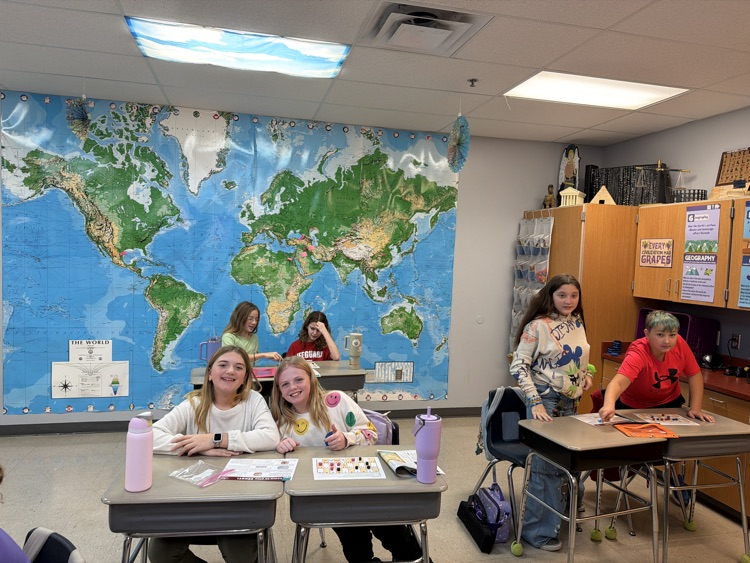sixth graders sitting at gray school desks. a world map hangs on the wall behind them.