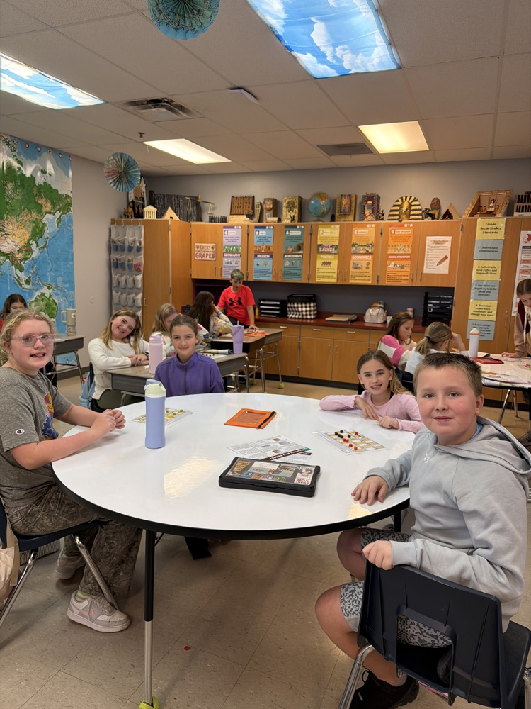 sixth graders sitting at a gray circle table, a row of wall cabinets and shelving is on the wall behind them