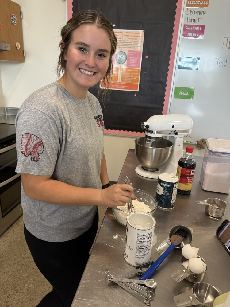 girl wearing gray t shirt and black pants, stirring flour in a glass bowl using a whisk