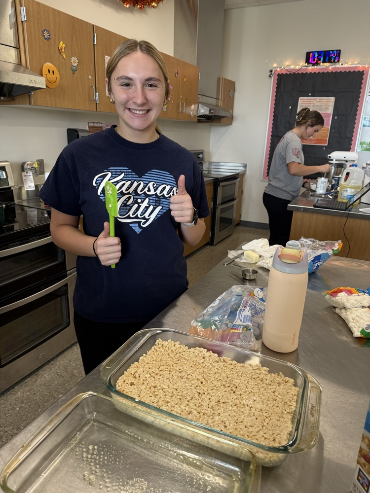 girl wearing blue t shirt and blue pants, giving a thumbs up with her left hand, her right hand holds a green spatula