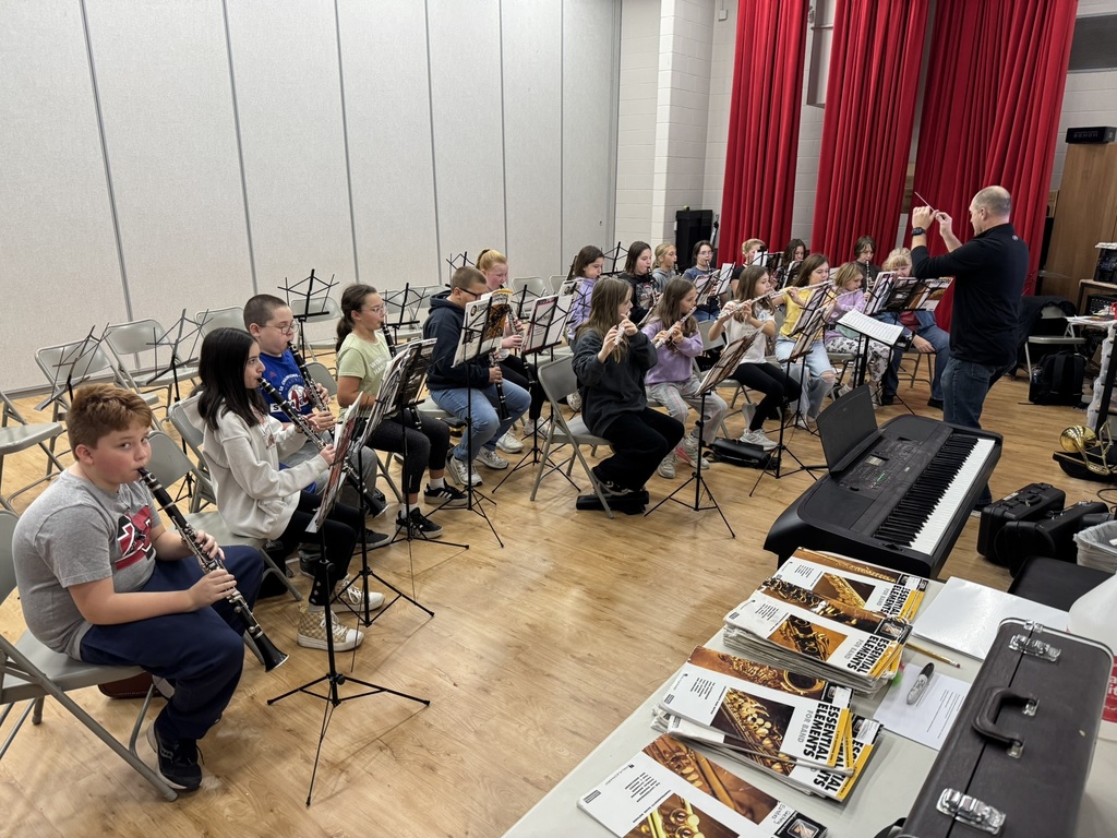 fifth grade students sitting in gray folding chairs playing flutes and clarinets; a man in jeans and a black shirt stands at the front of the room conducting