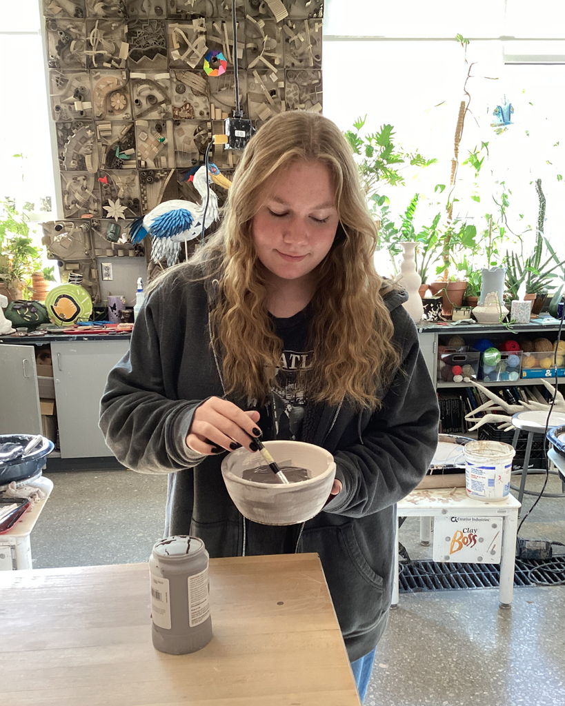 Hannah Turner wearing jeans and a dark gray sweatshirt. She holds a white ramen bowl in her left hand and glazes it with a gray liquid using a  brush in her right hand