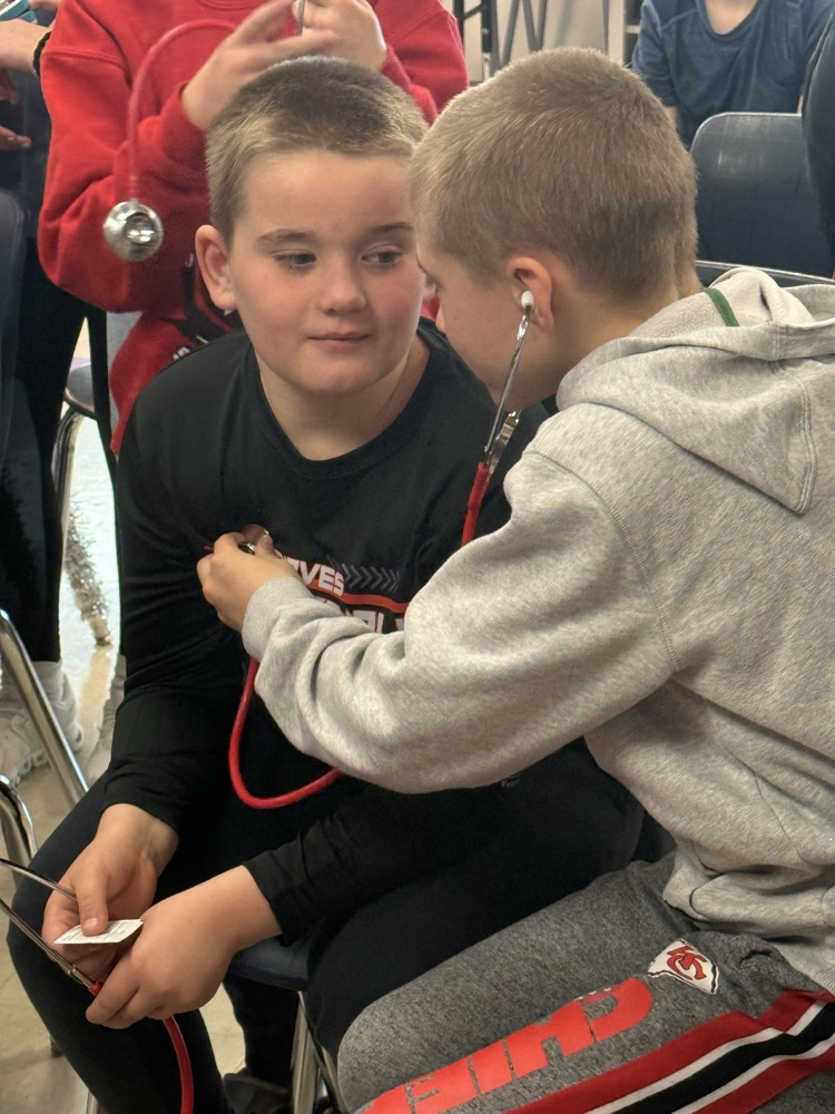 a fifth grade boy wearing a stethoscope listens to the heart of a fellow male classmate