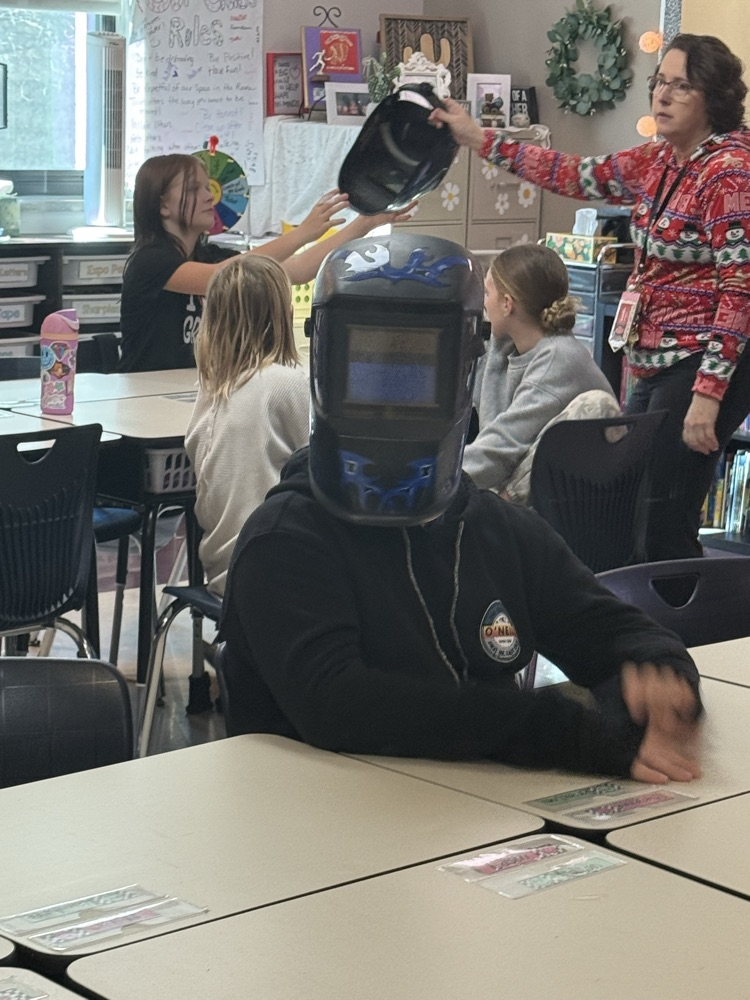 fifth grade boy sitting at a classroom desk tries on a black and blue welding helmet