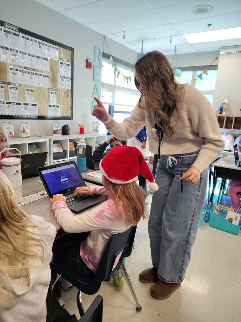 Madison wearing a cream sweater, jeans, and brown shoes, standing behind a girl sitting at a desk on a computer