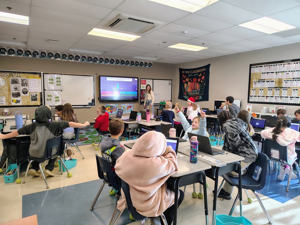 Madison wearing a cream sweater, jeans, and brown shoes, standing at the front of a classroom talking to second grade students, who are sitting in desks