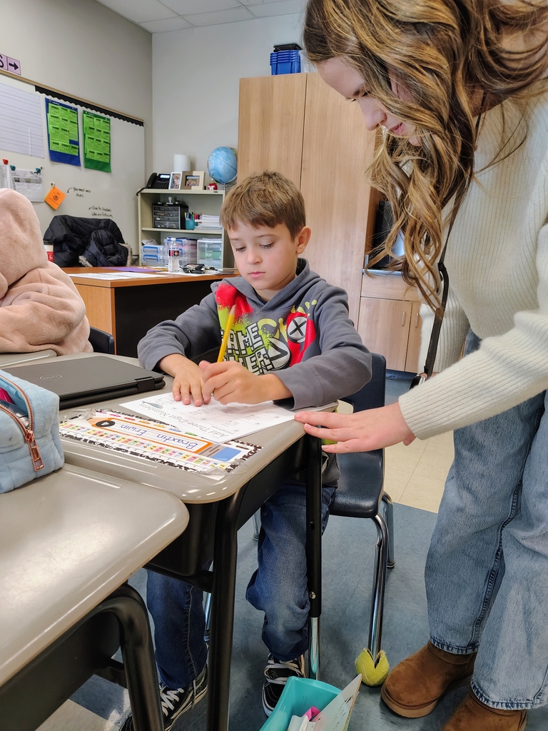 Madison wearing a cream sweater, jeans, and brown shoes, standing beside a boy sitting at a desk writing with a pencil