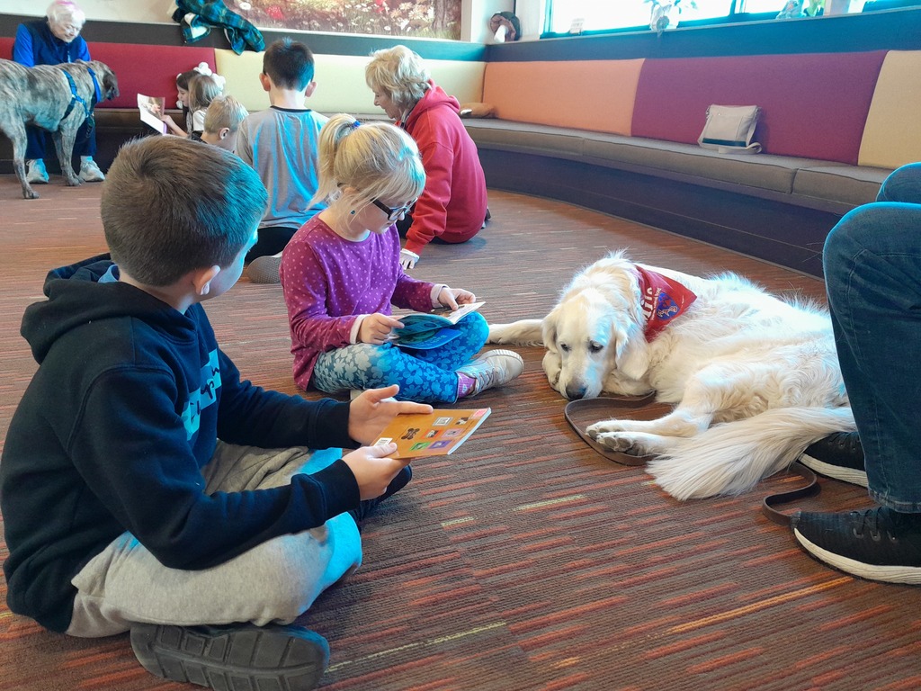 a girl and a boy reading paperback books to a white dog wearing a red bandanna