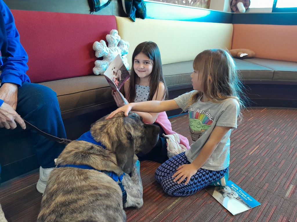 two girls reading a paperback book to a brown dog wearing a blue leash