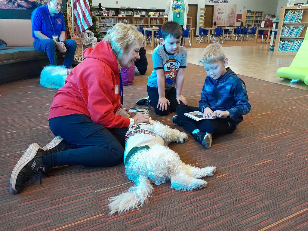 two boys reading a paperback book to a white dog laying on brown carpet