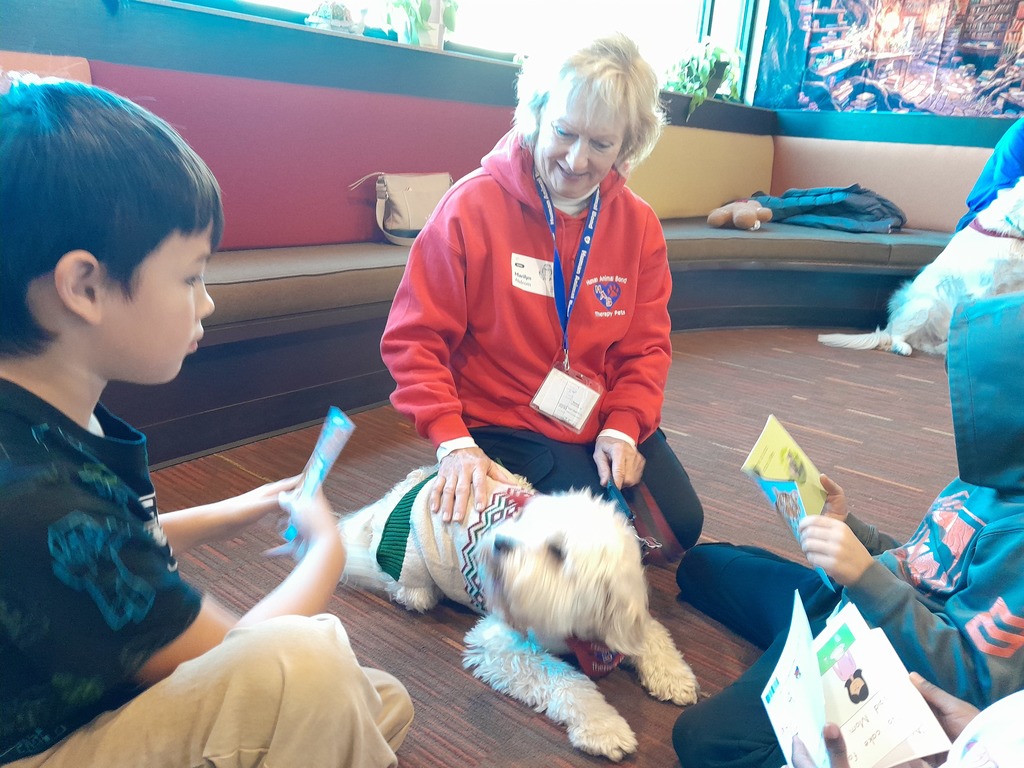 two fifth grade boys reading paperback books to a white dog laying on brown carper