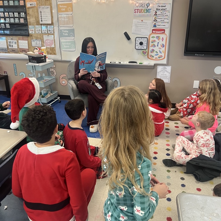 Elementary students dressed in festive holiday pajamas sit on a carpet, listening to a female adult read a book aloud