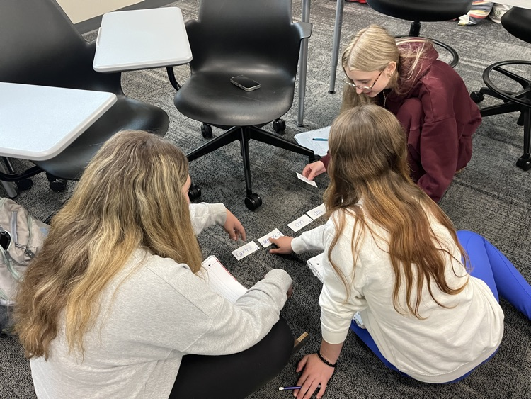 Three female students are sitting on the floor and are discussing the vocabulary cards that they sequenced to create a sentence from our class novel, El capibara con botas.