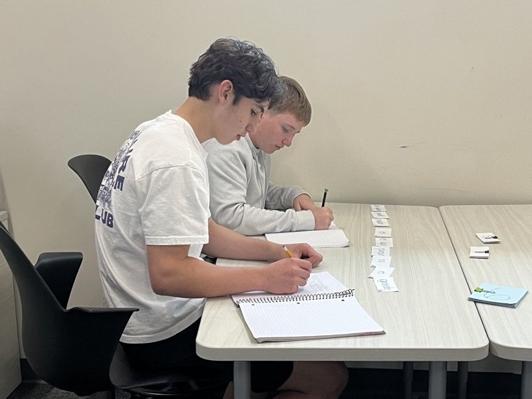 Two male students are sitting at a table and are copying their sentence that they sequenced using the vocabulary cards from our class novel, El capibara con botas.