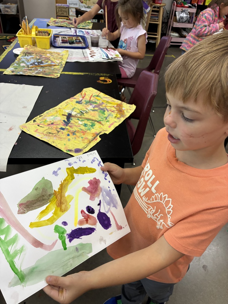 Kindergarten student showing his painting of lines and colors. Girl students painting in the background.