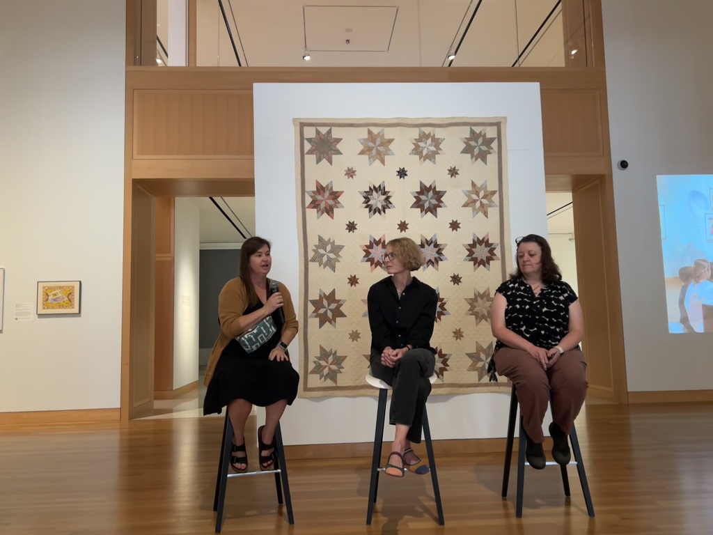 Teachers sitting on stools in front of a quilt in a museum. 