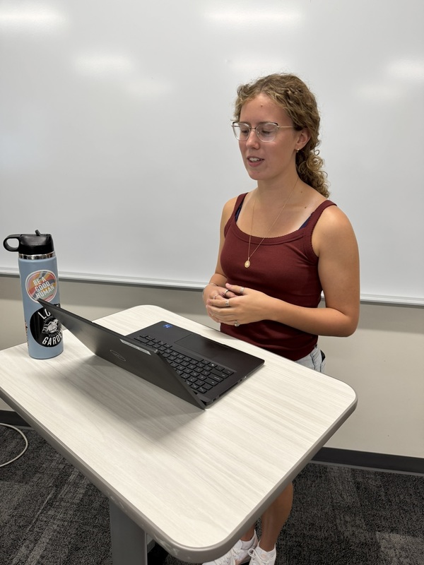 student wearing red tank top, standing at a rolling desk with her open laptop
