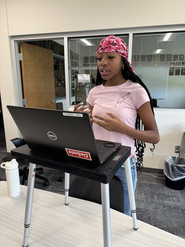 student wearing pink shirt and scarf standing before an open laptop sitting on a portable podium