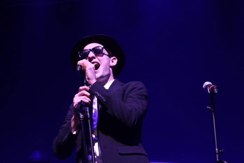 wesley williams wearing sunglasses and a black hat and suit, in Doobie Brothers style, singing into a microphone