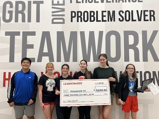 students holding a large white check in front of a word collage wall