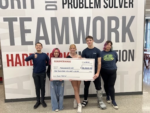 students holding a large white check in front of a word collage wall