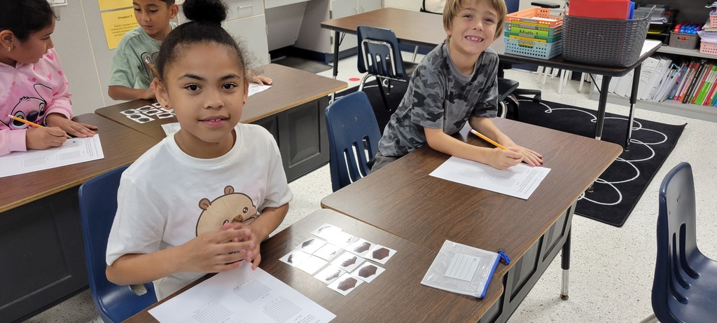 Two children, one holding a pencil, sit at desks in a classroom, facing papers and writing materials.