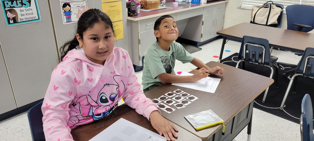 Two students sit at desks in a classroom. One wears a pink hoodie, the other a green shirt.