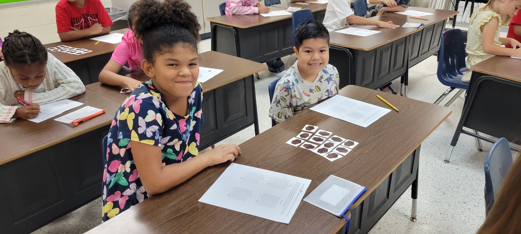 Children sitting at desks in a classroom, engaged in writing. Some have pens and papers on desks.