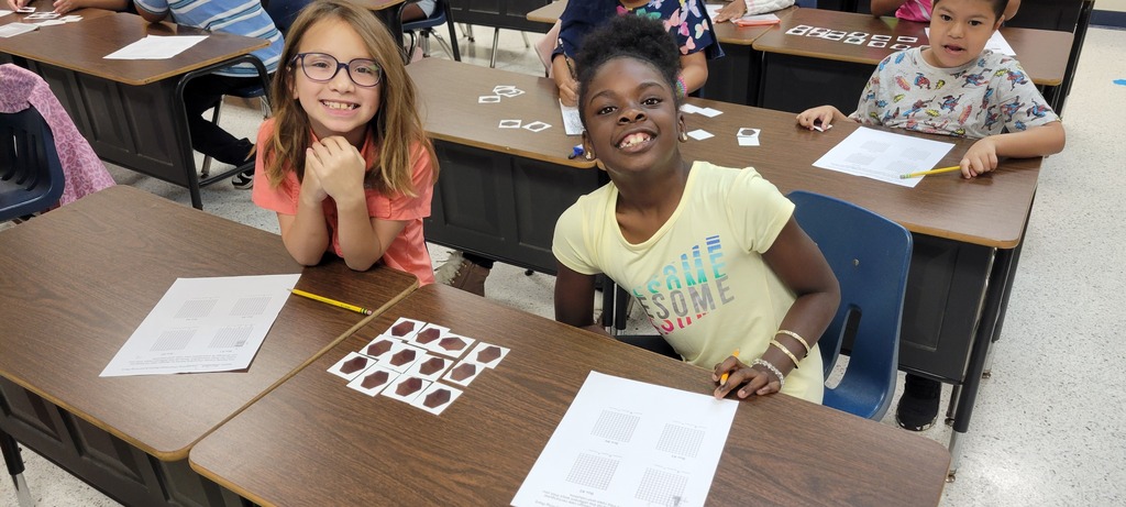 Four children sit at desks in a classroom. Two children smile at the camera. Papers, pencils, and puzzles on the desks.