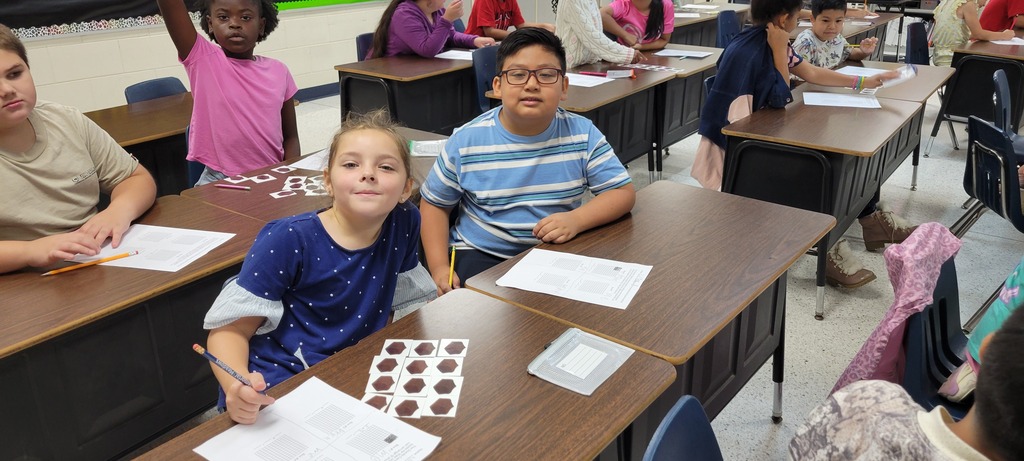 A classroom with students seated at desks. Papers and pencils are on the desks. One student is raising a hand.