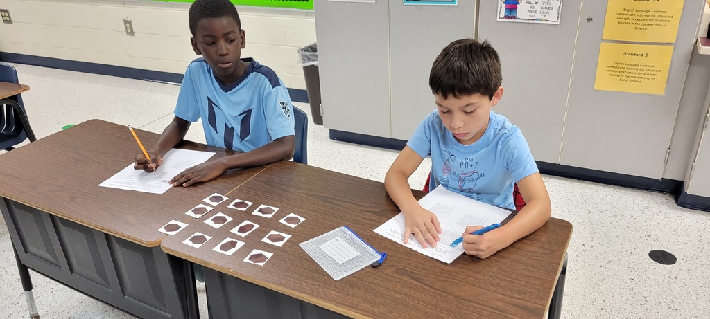 Two boys sit at a desk, each with paper and pencil, writing. One has a blue shirt and the other has a yellow shirt.