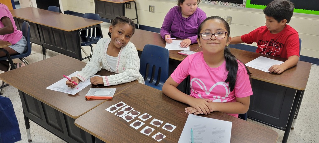Children in a classroom seated at desks, each writing with pens on paper, facing a blackboard.