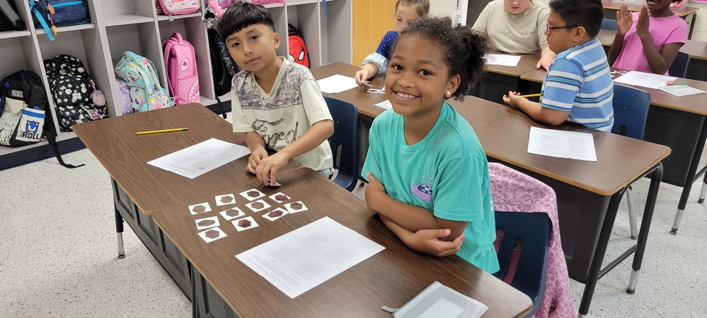 Children sit at desks in a classroom, working with papers. A child wears a blue shirt.