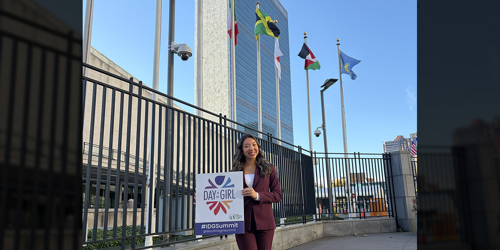 Photo of Sofia posing with a flyer avertising the summit, with the flags of various countries flying behind her