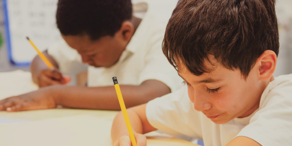 wo students seated at a classroom table, writing with pencils on paper during a lesson.